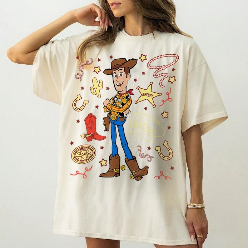May include: Cream-colored t-shirt featuring a cartoon illustration of Woody from Toy Story. The design includes Woody in his cowboy attire, surrounded by western-themed graphics like stars, a sheriff badge, and a horseshoe. The shirt is a casual, oversized fit.