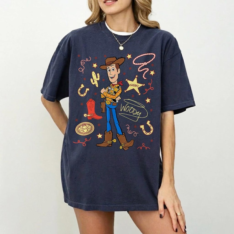 May include: Navy blue t-shirt featuring a graphic of Woody from Toy Story. The design includes a sheriff's badge, cowboy boots, and a cactus. The word "Woody" is written in a playful font. The shirt is a relaxed fit.