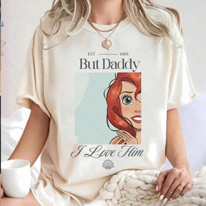 May include: A cream-coloured t-shirt featuring the text "But Daddy" above a cartoon image of a red-haired character and the words "I Love Him" below. The shirt has short sleeves and a relaxed fit.