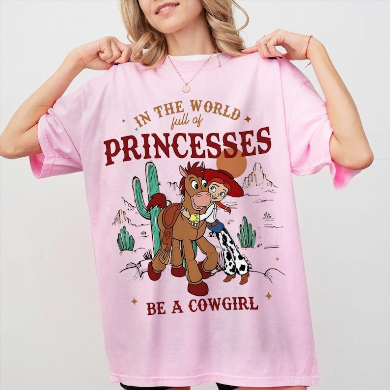 以下が含まれることがあります： 「IN THE WORLD full of PRINCESSES BE A COWGIRL」の文字と、カウガールと馬のイラストが描かれたピンク色のTシャツ。 短い袖と丸い襟が特徴です。