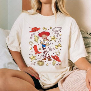 Peut inclure: T-shirt cr&egrave;me avec une illustration de dessin anim&eacute; d'une cowgirl nomm&eacute;e "Jessie". Le motif comprend un chapeau rouge, des bottes, des &eacute;toiles et un lasso. Le t-shirt est de coupe ample.