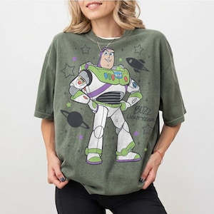 Puede incluir: Camiseta verde oliva con un dise&ntilde;o de Buzz Lightyear de Toy Story. El dise&ntilde;o incluye a Buzz con su traje espacial, estrellas, un cohete y el texto "Buzz Lightyear". La camiseta tiene mangas cortas y un corte holgado.