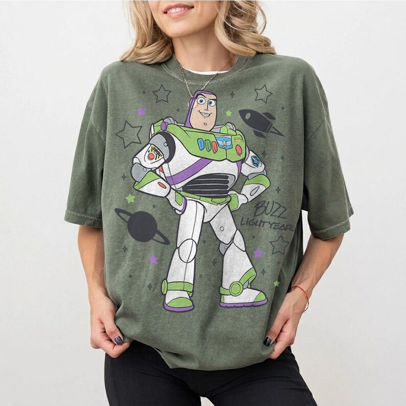 Puede incluir: Camiseta verde oliva con un dise&ntilde;o de Buzz Lightyear de Toy Story. El dise&ntilde;o incluye a Buzz con su traje espacial, estrellas, un cohete y el texto "Buzz Lightyear". La camiseta tiene mangas cortas y un corte holgado.