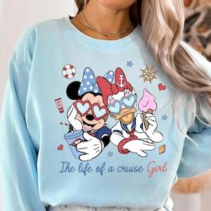 May include: Light blue sweatshirt featuring Minnie Mouse and Daisy Duck in heart-shaped sunglasses, holding drinks and ice cream. The text "The life of a cruise Girl" is printed below the characters. Nautical themed design.