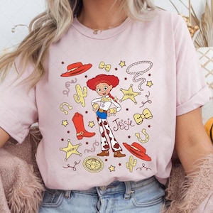 Peut inclure: T-shirt rose clair avec une illustration de dessin anim&eacute; de Jessie de Toy Story. Le design comprend un chapeau rouge, des bottes et le nom "Jessie". Des &eacute;l&eacute;ments suppl&eacute;mentaires incluent des &eacute;toiles, un lasso et un badge de sh&eacute;rif.