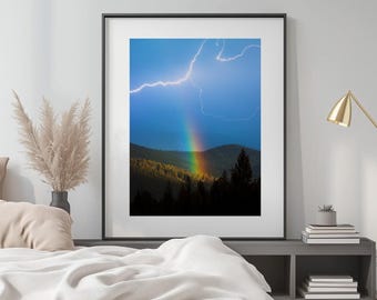 May include: Framed photograph showing a rainbow over a forest and mountains, with a lightning bolt in the sky. The rainbow displays a full spectrum of colours. The photograph is displayed in a black frame.
