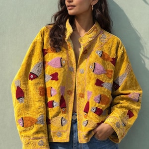 May include: A yellow velvet jacket with colorful embroidered fish designs. The jacket features a mandarin collar and is open, revealing the wearer's clothing. The fish are in shades of red, pink, orange, and gray. The jacket is paired with blue jeans.