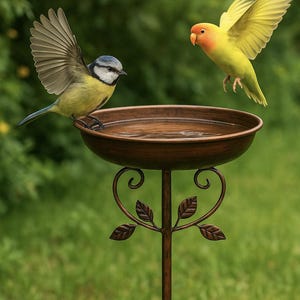 Puede incluir: Un bebedero de bronce con un pájaro azul y amarillo posado en el borde. Otro pájaro amarillo está en vuelo, con las alas extendidas. El bebedero tiene un tallo decorativo con detalles de hojas, sobre un fondo verde borroso.