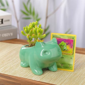 May include: A light green ceramic Bulbasaur planter with a small succulent plant inside. A Bulbasaur trading card is next to the planter. The planter is on a light brown surface.