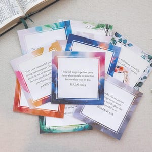 May include: A collection of square cards with watercolor borders and inspirational quotes from the Bible. The cards feature colorful designs and text, with a partially visible open book in the background.