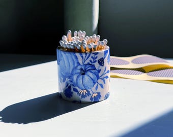 May include: A ceramic match holder with a blue and white floral design, filled with matches. The holder features a pumpkin and berry pattern. The matches have white tips. A few yellow and purple stickers are in the background.