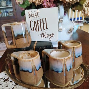 May include: Four ceramic coffee mugs with a unique design, featuring a brown, blue, and beige glaze. The mugs are displayed on a decorative tray, with a sign that reads "First I drink the coffee then I do the things."