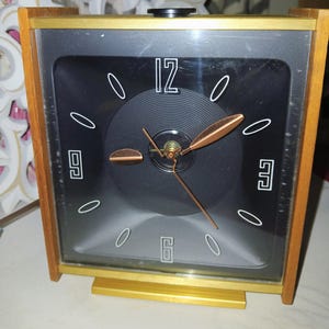 May include: A vintage square clock with a black face, white numerals, and copper-colored hands. The clock has a wooden frame with gold accents and a small black knob on top. The clock measures approximately 6 inches wide and 6 inches tall.