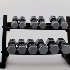 K&ouml;nnte beinhalten: Ein schwarzes Hantelregal mit mehreren grauen, sechseckigen Hanteln. Die Gewichte sind von 23 bis 45 kg angegeben. Das Regal hat zwei Ebenen und besteht aus schwarzem Metall, vor einem wei&szlig;en Hintergrund.