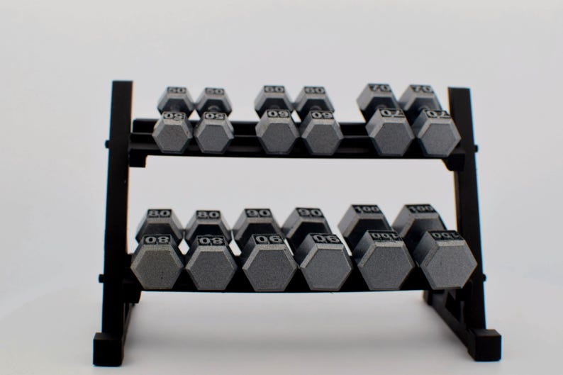 K&ouml;nnte beinhalten: Ein schwarzes Hantelregal mit mehreren grauen, sechseckigen Hanteln. Die Gewichte sind von 23 bis 45 kg angegeben. Das Regal hat zwei Ebenen und besteht aus schwarzem Metall, vor einem wei&szlig;en Hintergrund.