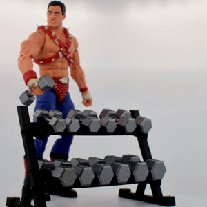 K&ouml;nnte beinhalten: Ein schwarzes Hantelregal gef&uuml;llt mit grauen Hanteln. Eine muskul&ouml;se Actionfigur in Wrestling-Kleidung h&auml;lt eine Hantel. Die Figur tr&auml;gt rote und blaue Kleidung mit Armb&auml;ndern und einem Nietengeschirr.