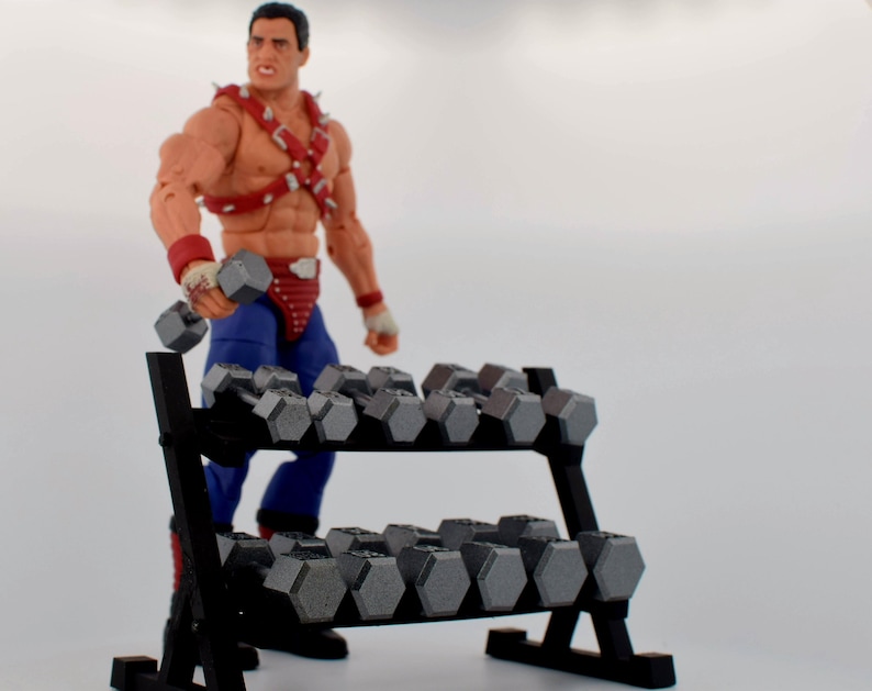 K&ouml;nnte beinhalten: Ein schwarzes Hantelregal gef&uuml;llt mit grauen Hanteln. Eine muskul&ouml;se Actionfigur in Wrestling-Kleidung h&auml;lt eine Hantel. Die Figur tr&auml;gt rote und blaue Kleidung mit Armb&auml;ndern und einem Nietengeschirr.