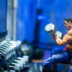 K&ouml;nnte beinhalten: Figur einer muskul&ouml;sen Figur, die in einem Fitnessstudio zwei silberne Kurzhanteln hebt. Er tr&auml;gt einen roten Gurt mit silbernen Stacheln und eine blaue Hose. Im Hintergrund befindet sich ein Hantelst&auml;nder.