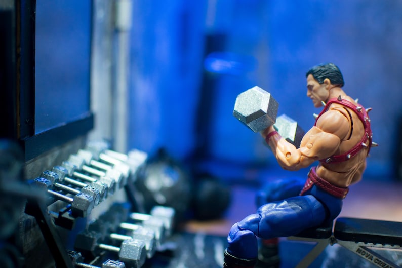 K&ouml;nnte beinhalten: Figur einer muskul&ouml;sen Figur, die in einem Fitnessstudio zwei silberne Kurzhanteln hebt. Er tr&auml;gt einen roten Gurt mit silbernen Stacheln und eine blaue Hose. Im Hintergrund befindet sich ein Hantelst&auml;nder.