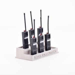 Puede incluir: Un conjunto de walkie-talkies negros con antenas, dispuestos en un soporte gris claro. Cada dispositivo tiene un teclado y detalles rojos. El soporte lleva el texto "NOVA CREATIONS".