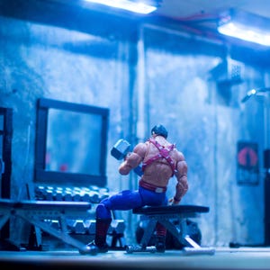 K&ouml;nnte beinhalten: Eine Miniatur-Actionfigur in einem Fitnessstudio, die eine Hantel hebt. Die Figur tr&auml;gt eine blaue Hose, ein rotes Geschirr und einen schwarzen Helm. Das Fitnessstudio hat Gewichte, B&auml;nke und ein Trainingsger&auml;t. Die Szene ist blau beleuchtet.