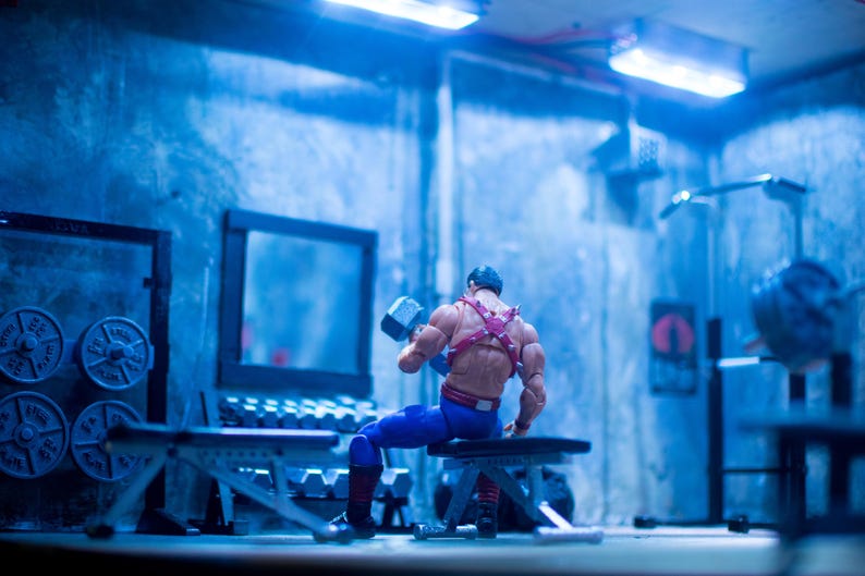 K&ouml;nnte beinhalten: Eine Miniatur-Actionfigur in einem Fitnessstudio, die eine Hantel hebt. Die Figur tr&auml;gt eine blaue Hose, ein rotes Geschirr und einen schwarzen Helm. Das Fitnessstudio hat Gewichte, B&auml;nke und ein Trainingsger&auml;t. Die Szene ist blau beleuchtet.