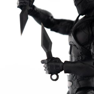 May include: A black action figure of a character in a tactical suit, holding two bladed weapons. The figure is in a dynamic pose, with one weapon held high and the other at the ready. The figure is set against a white background.