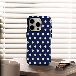 May include: A navy blue phone case with a white polka dot pattern. The case has a glossy finish and a camera cutout. A brown leather watch with a silver buckle is next to the phone case.