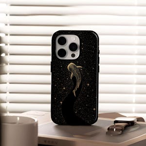 May include: Black phone case with a gold whale shark design. The case has a starry night sky pattern with a whale shark swimming upwards. The case has a camera cutout and is sitting on a book.