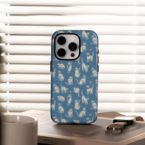 May include: A blue phone case featuring a repeating pattern of white cat illustrations in various poses. The case has a black border and a camera cutout. The phone case is sitting on a book next to a watch.