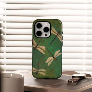 Peut inclure: Coque de téléphone verte avec un motif de libellules. L'étui a un fond vert foncé avec plusieurs libellules dans des tons de beige et de marron. Les libellules sont détaillées avec des ailes et des corps visibles. L'étui est conçu pour un smartphone.