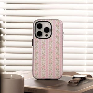 Pode incluir: Uma capa de smartphone com um padrão floral de rosas cor-de-rosa e folhas verdes sobre um fundo branco. A capa tem uma borda preta e um recorte para a câmera. A capa do telefone está em uma mesa com um relógio e um livro.
