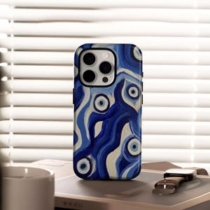 May include: A mobile phone case with a blue and white abstract design featuring eye-like shapes. The case has a black border and a camera cutout. The design incorporates various shades of blue and white, creating a wavy pattern. The case is on a table with a watch and a cup.