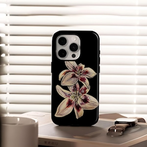 May include: Black phone case featuring a watercolor illustration of two lilies. The flowers have white petals with maroon and yellow accents. The case is shown on a book with a watch.