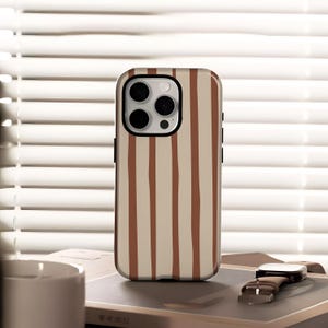 May include: A phone case with a vertical striped pattern in beige and brown. The case has a black border around the camera and a black frame. The phone case is resting on a book next to a watch.