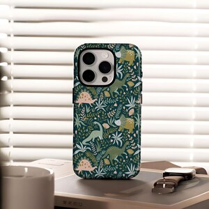 Puede incluir: Una funda de teléfono verde oscuro con un patrón repetido de dinosaurios, plantas y flores en verde, rosa y amarillo. La funda tiene un borde negro y un recorte para la cámara.