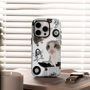 May include: A white phone case featuring a collage of cat-themed illustrations. The design includes a cat on a skateboard, a cat outline, an 8-ball, dice, and other playful graphics. The background has a newspaper print.