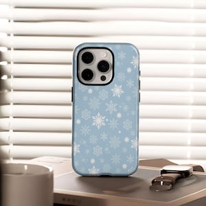 Peut inclure: Coque de téléphone bleu clair ornée de flocons de neige blancs. Le boîtier a un bord noir et une découpe pour l'appareil photo. Les flocons de neige sont de tailles variables. La coque est posée sur un livre avec une montre à côté.