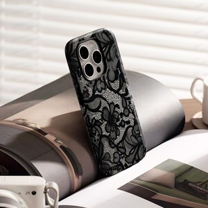 May include: Black phone case with a delicate lace pattern. The case features a floral design with intricate details, offering a stylish and protective accessory. The case is designed to fit a smartphone.