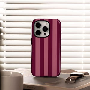 May include: A burgundy and pink striped phone case. The case has a glossy finish and a black border. The phone case is resting on a book next to a watch with a brown leather band.