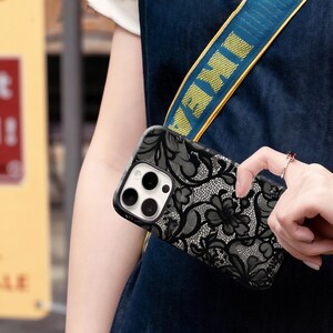 May include: Black and gray floral lace phone case. The case has a black border and a camera cutout. The phone case is being held by a person wearing a blue denim top. A blue and yellow strap with the word "IKEA" is visible.