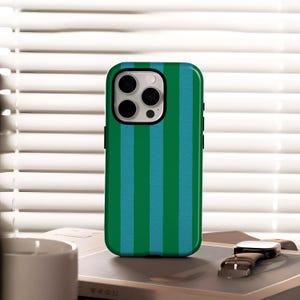 Può includere: Custodia per telefono a righe verdi e blu con modulo fotocamera nero. La custodia è appoggiata su un libro accanto a un orologio con cinturino in pelle marrone. Lo sfondo presenta una persiana bianca.
