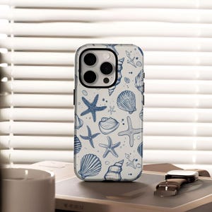 Puede incluir: Funda de teléfono blanca con un diseño de conchas marinas y estrellas de mar azules. La funda tiene un recorte para la cámara y está sobre un libro. Un reloj con una correa de cuero marrón está al lado del teléfono.
