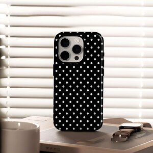 May include: Black smartphone case with a white polka dot pattern. The case has a square shape with rounded corners and a camera cutout. The phone case is on a book next to a watch with a brown leather strap.