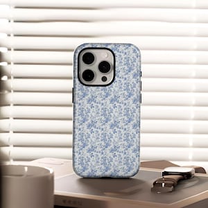 Puede incluir: Funda de teléfono con estampado floral en tonos azules y blancos. La funda está sobre una mesa con un reloj y una taza blanca. El fondo presenta persianas blancas.
