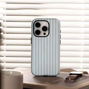 May include: A light blue and white striped phone case with a black border. The case has a camera cutout and is shown on a book with a watch and a white mug in the background.