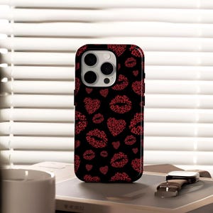 May include: Black phone case featuring a pattern of red leopard print lips and hearts. The case has a glossy finish and a camera cutout. The phone case is on a book.