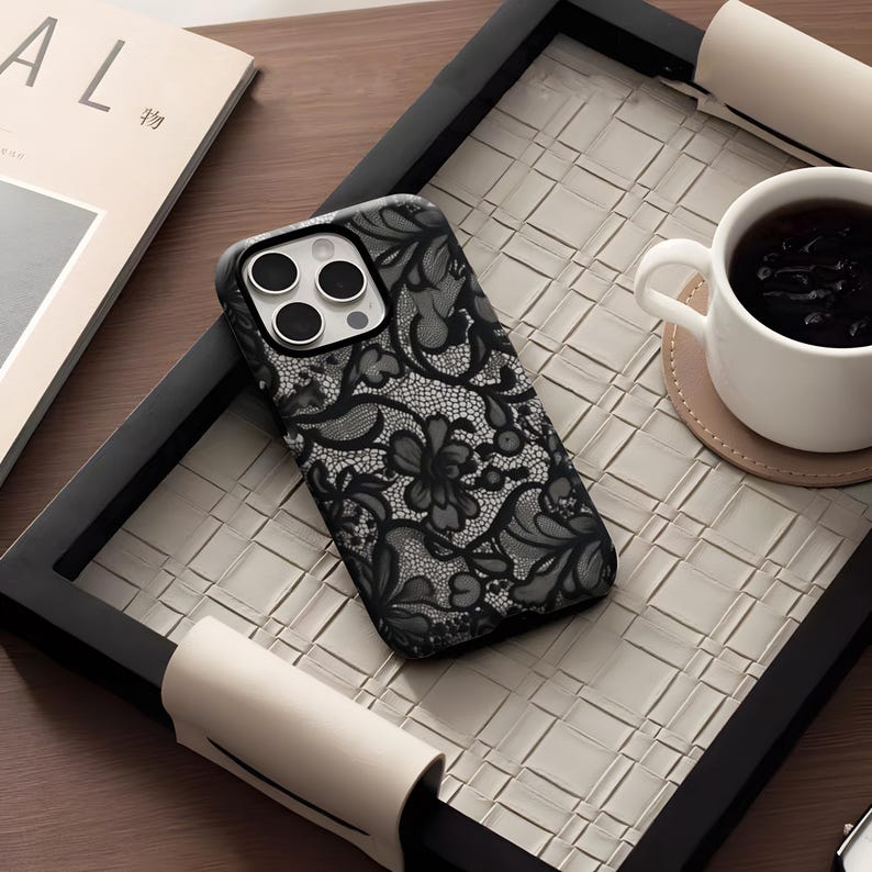 May include: A black phone case with a delicate lace pattern, resting on a woven tray. The case features a floral design and is next to a white teacup on a coaster. A magazine is visible in the background.