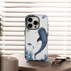 May include: White phone case featuring a blue whale shark illustration with sea creatures like turtles, seahorses, and coral. The case has a black camera ring. A brown watch is visible in the background.