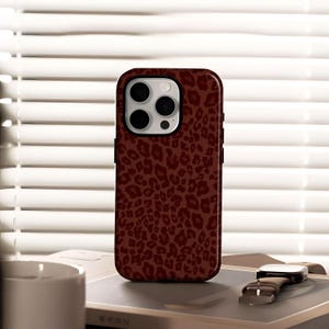 May include: A maroon phone case with a leopard print design. The case has a black border and a camera cutout. The phone case is sitting on a book next to a watch with a brown leather strap.